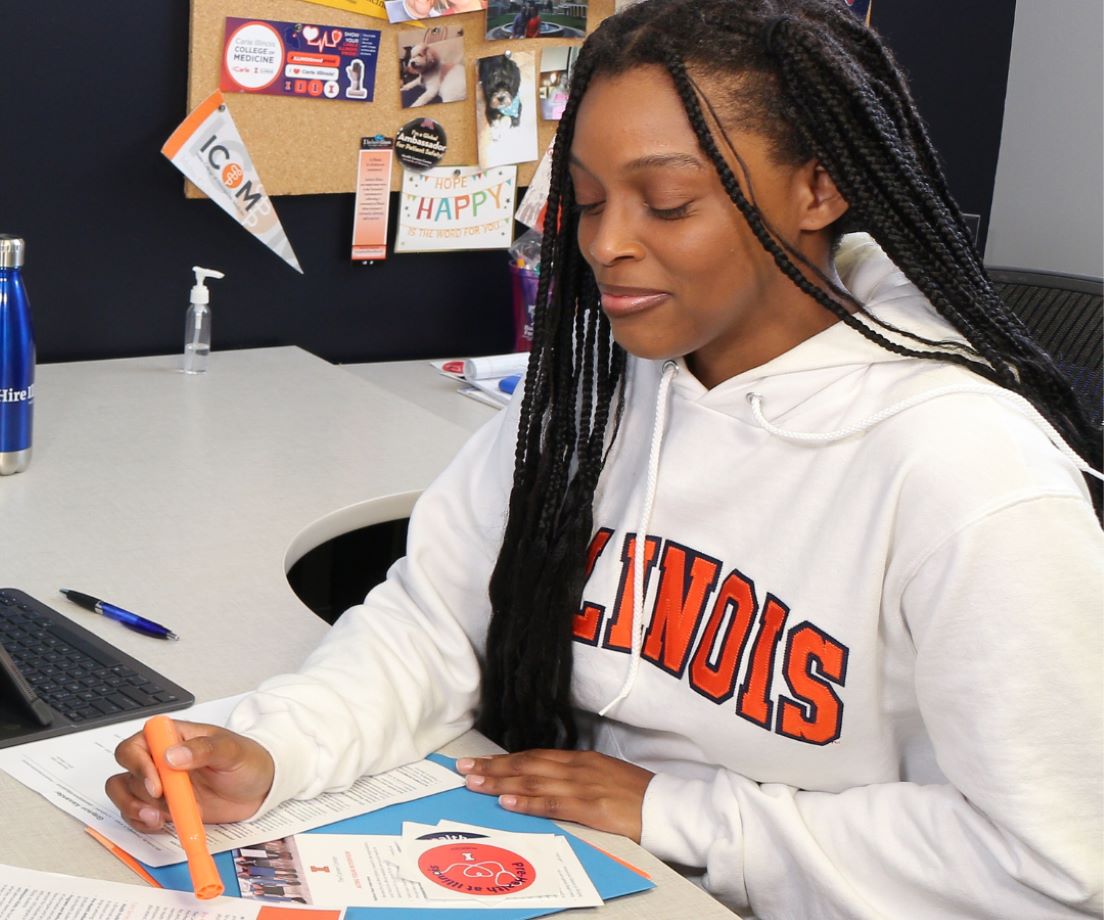 Pre-health ambassador wearing Illinois sweatshirt sits at a desk and points to information she is sharing