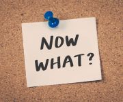 Small sticky note pinned to cork board reading "Now What?"