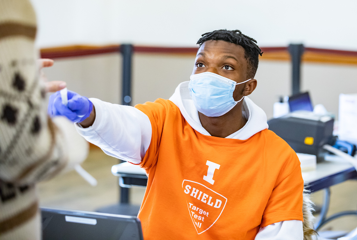 A student in a mask and gloves wearing an Illinois Shield shirt hands a plastic vial to someone for COVID testing