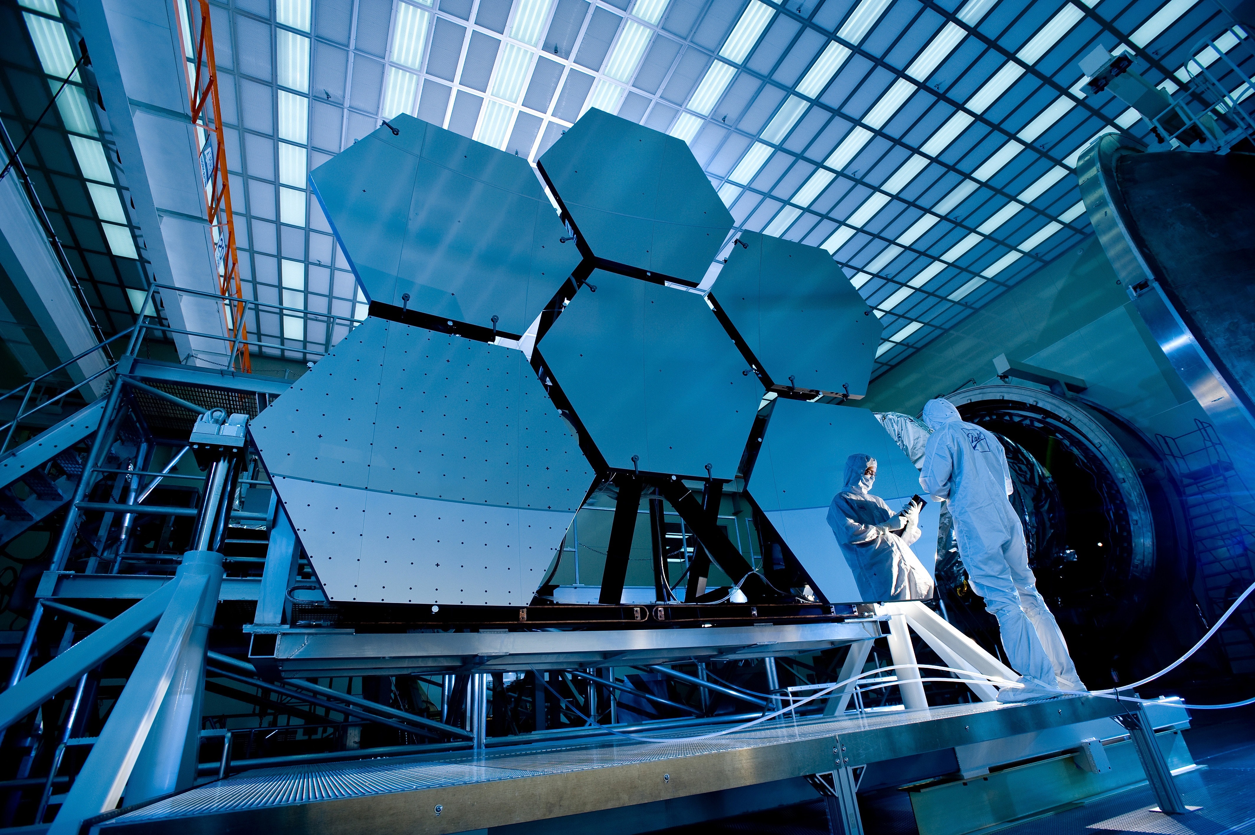 two people in white protective suits examine hexagonal solar panel array inside a closed warehouse environment