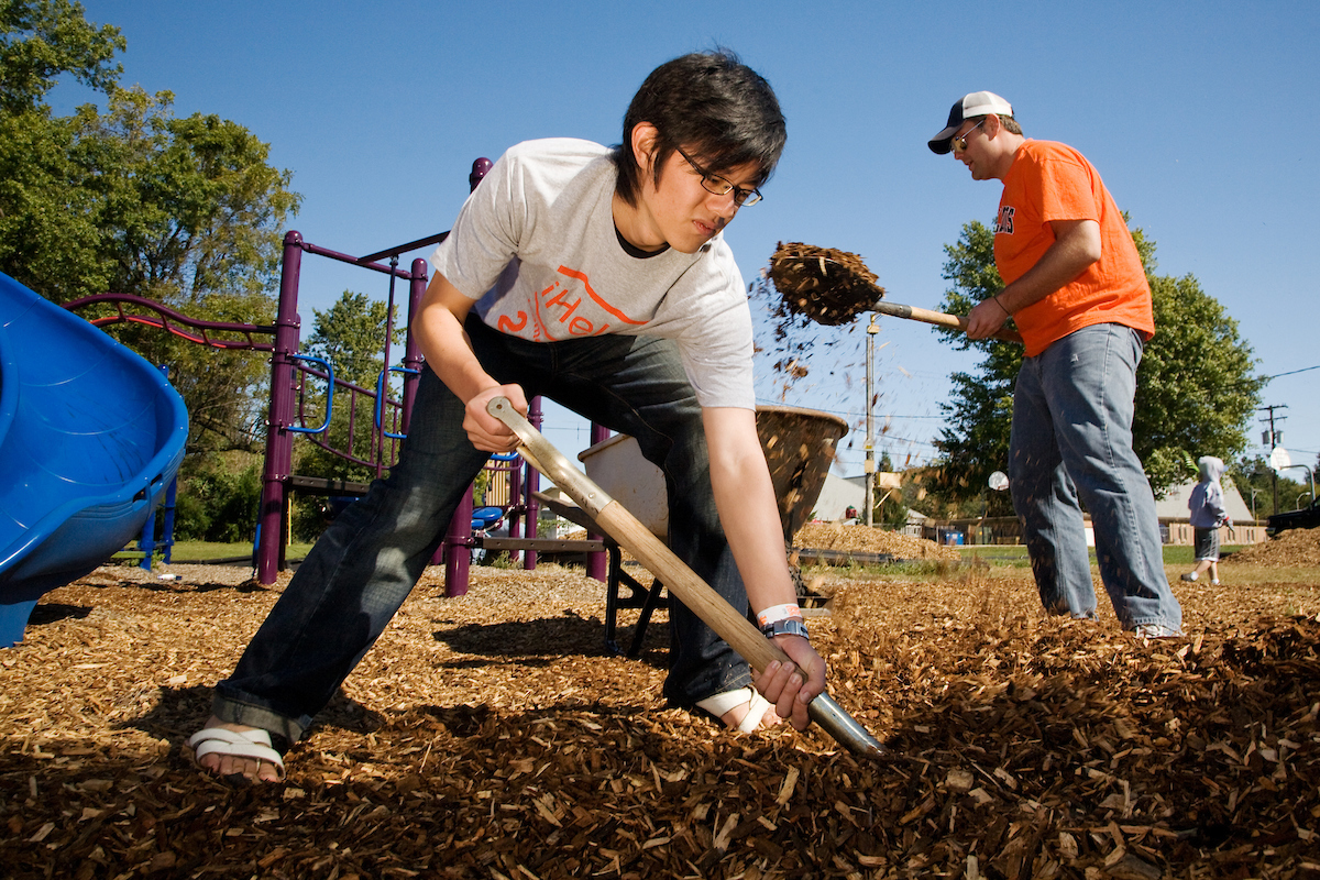 two student volunteers wearing iHelp shirts shovel mulch in a playground