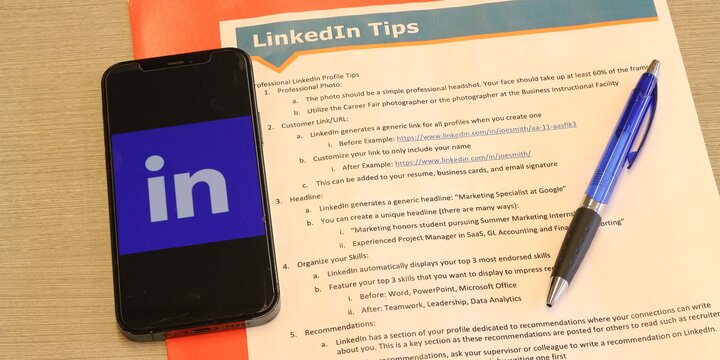 "LinkedIn Tips" instructional flyer lying on a desk next to a cellphone with LinkedIn logo on the screen, and a pen