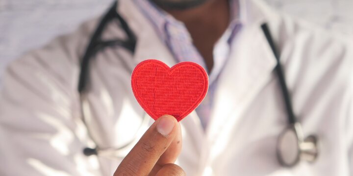 A person in a white doctor's coat and stethoscope holds out a cut-out of a red heart
