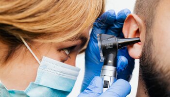 Woman wearing mask and gloves looking into bearded patient's ear with otoscope