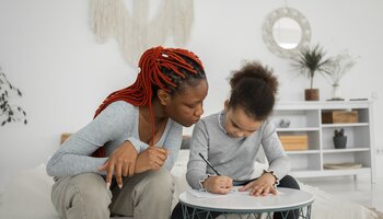 A woman with braided hair sits next to a younger child and watches as the child writes something at a small table